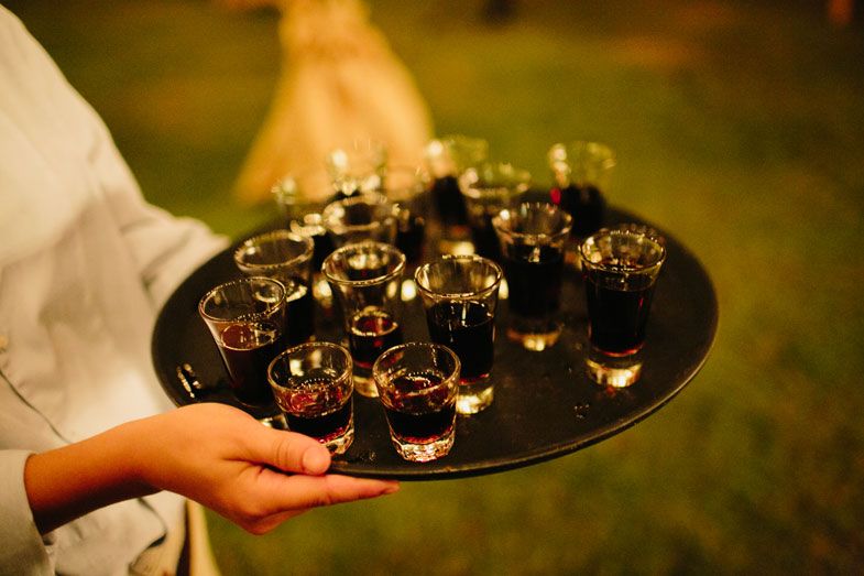 A server holds a black tray of dark beverages in small glasses at an evening event.