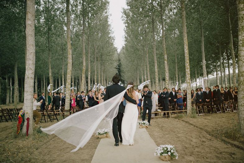 Wedding couple walking down aisle with bride's veil flowing in forest setting.