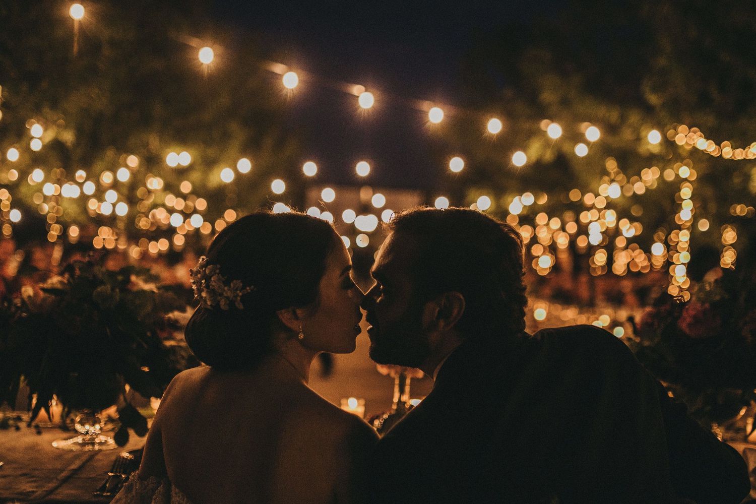 Romantic silhouette of couple beneath twinkling outdoor string lights at night.