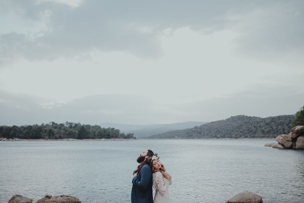A couple shares an intimate embrace on rocky shores with mountains and lake in the background.