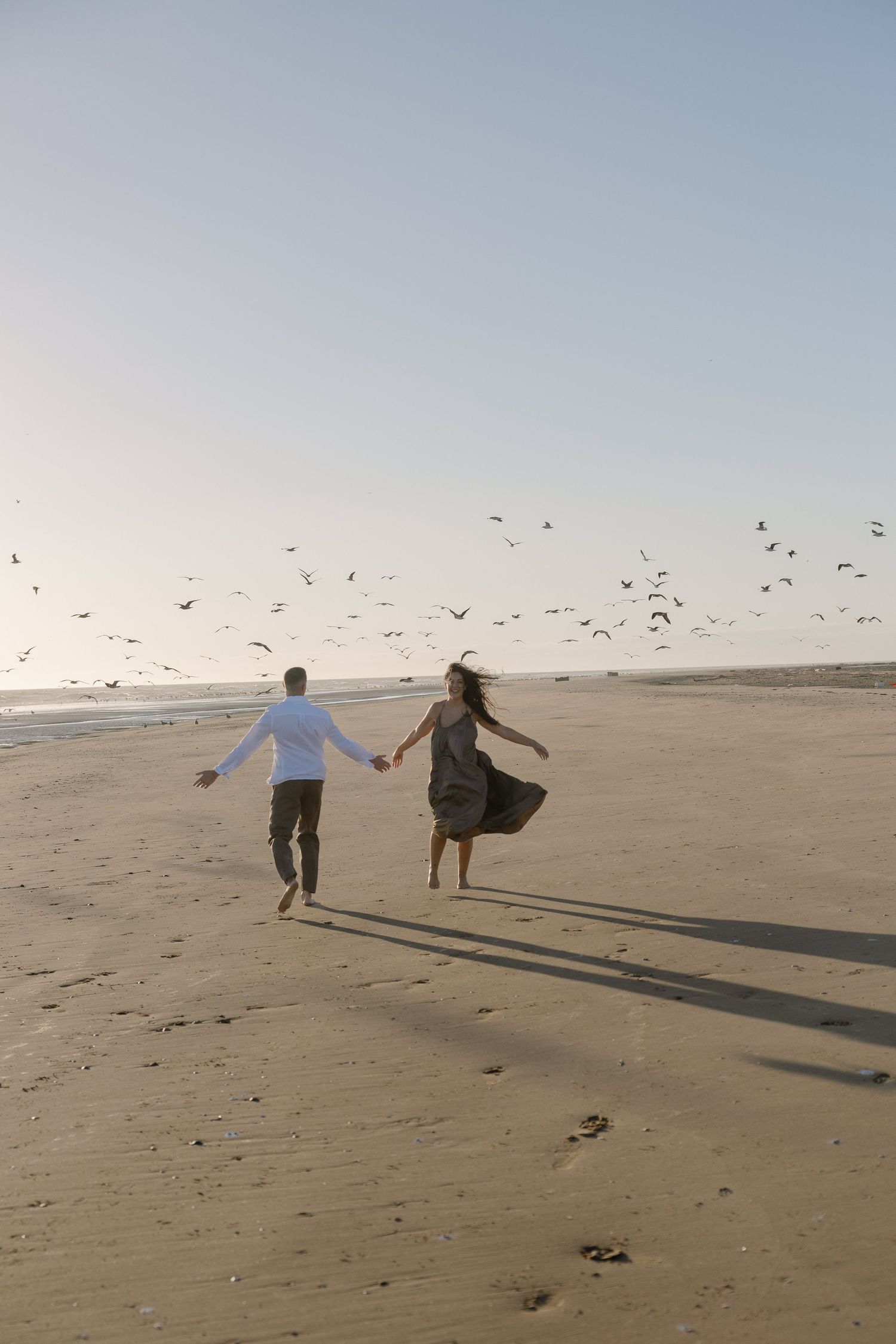Couple holding hands running on sandy beach with birds flying overhead in soft natural light.