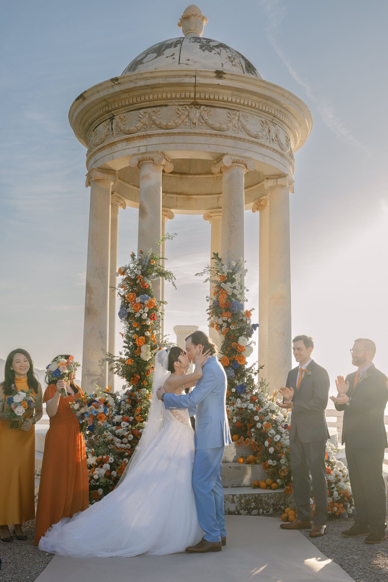 Bride and groom share first kiss at elegant stone rotunda altar adorned with orange and blue floral arrangements.