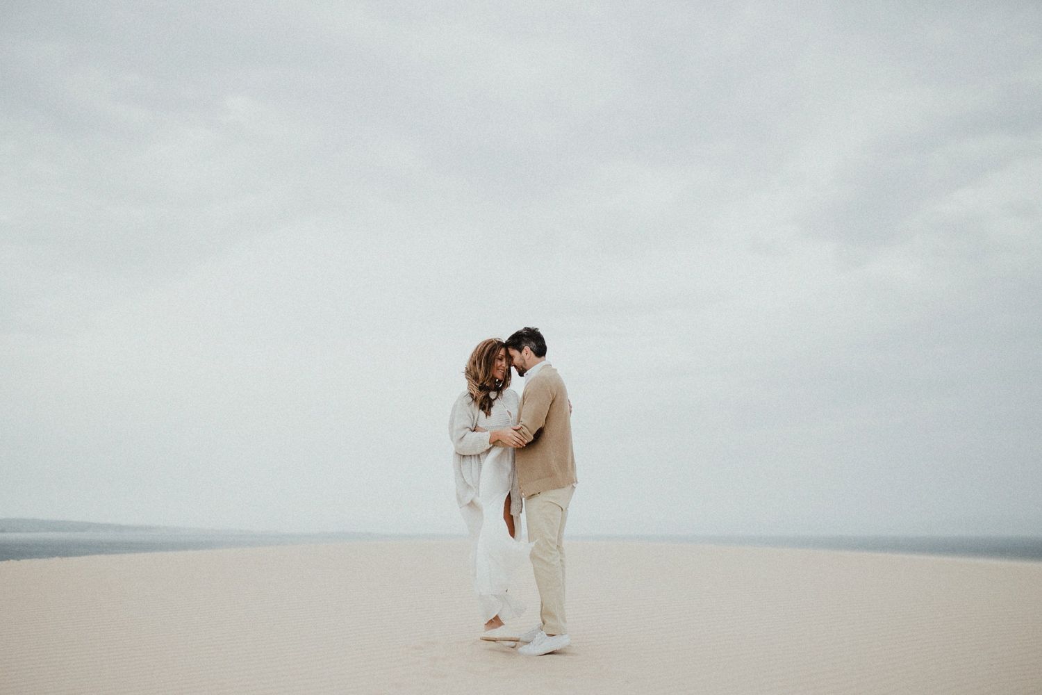Couple in light colored clothing sharing intimate moment on sandy dunes.