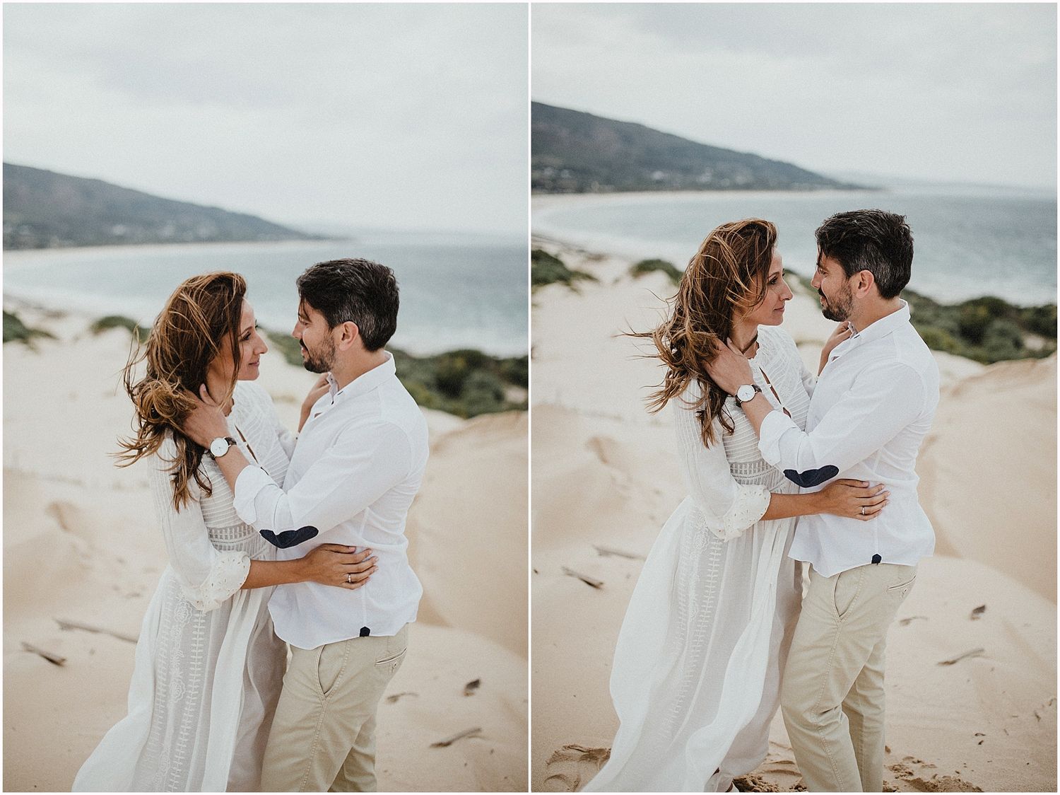 Romantic couple in white outfits embracing on a scenic beach with mountains in the background.