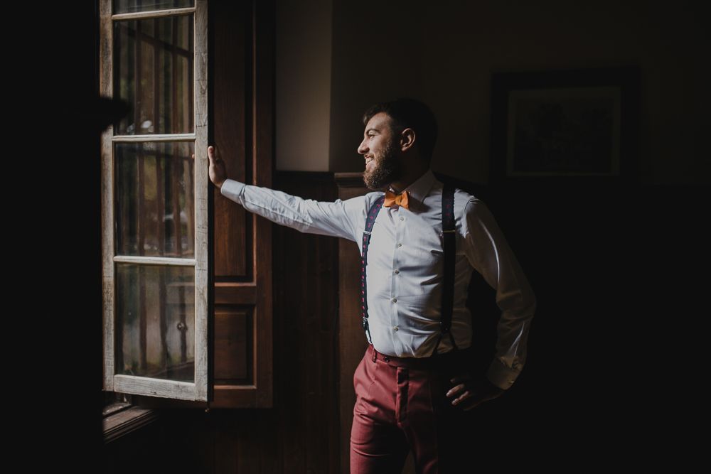 Person in formal attire poses by window with hand on hip and dramatic lighting.