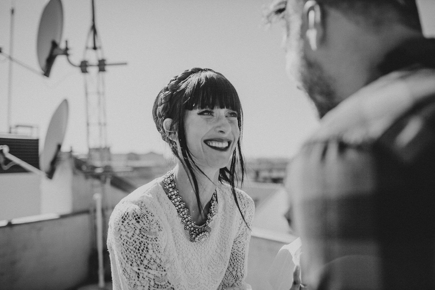 A woman in a white lace dress and statement necklace smiles during an outdoor rooftop ceremony.