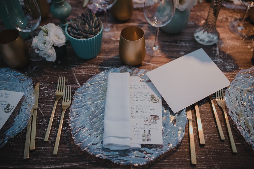 A rustic wedding table setting with gold cutlery, blue plates, and blank place cards on a wooden surface.