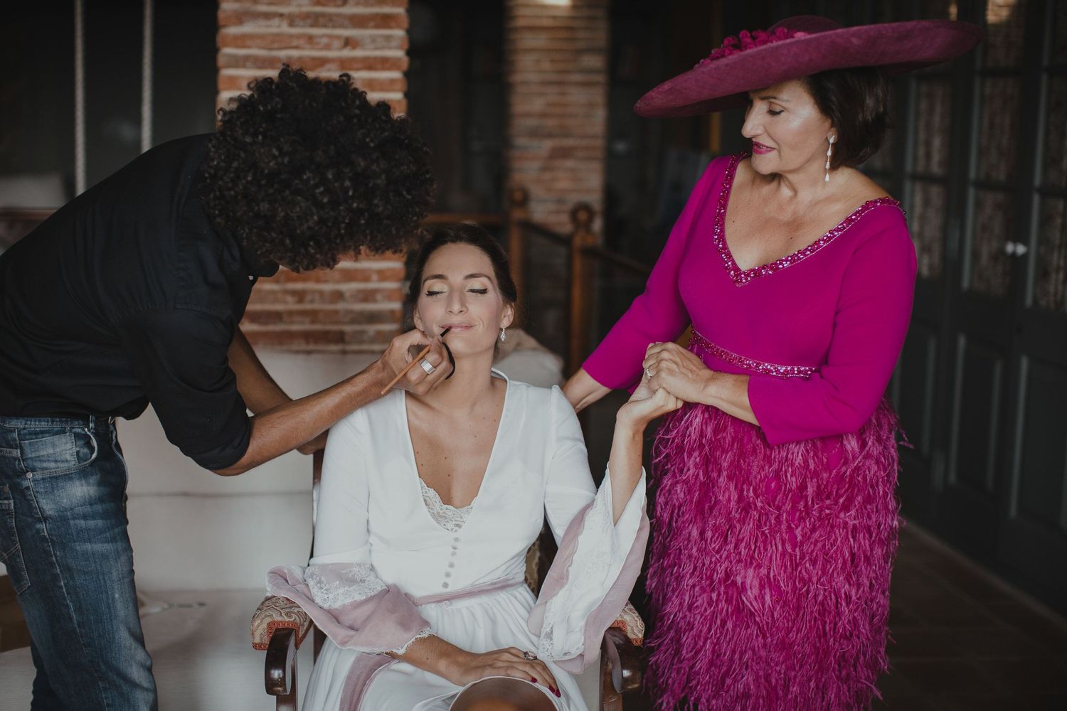 Wedding preparations with makeup artist and attendant in vibrant fuchsia dress helping with final touches.