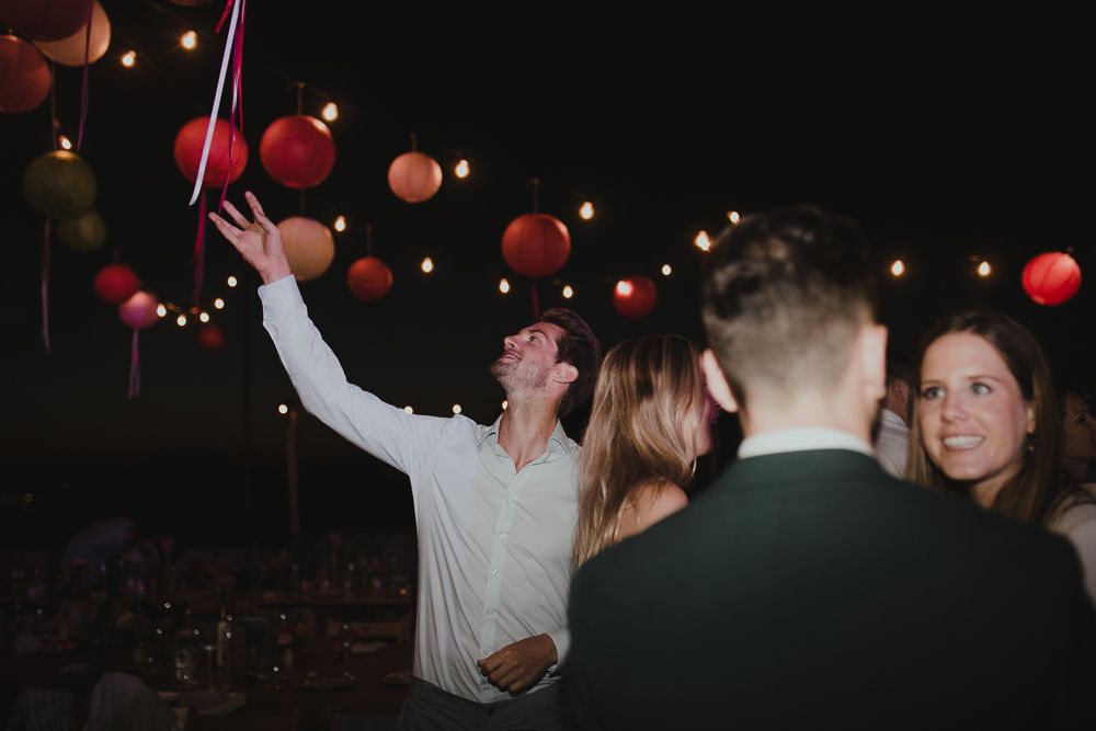 Wedding guests dancing and reaching for hanging lanterns at nighttime reception.