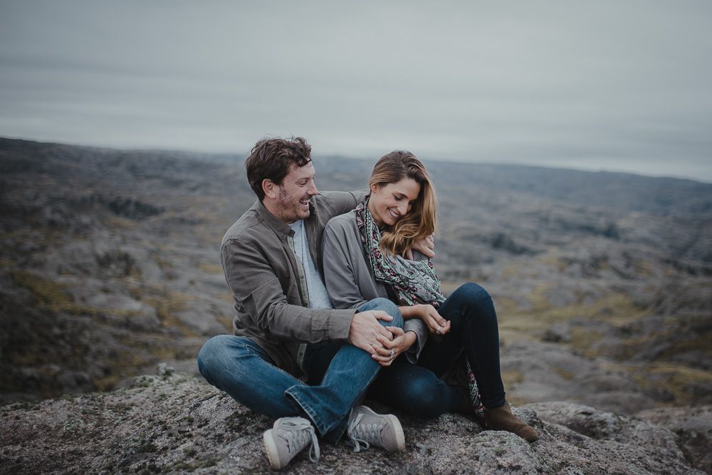 Couple in gray coats share intimate conversation against moody mountain landscape.