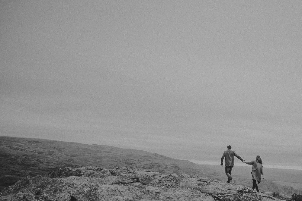 Two figures walk hand in hand across a rugged hilltop landscape against a moody sky.
