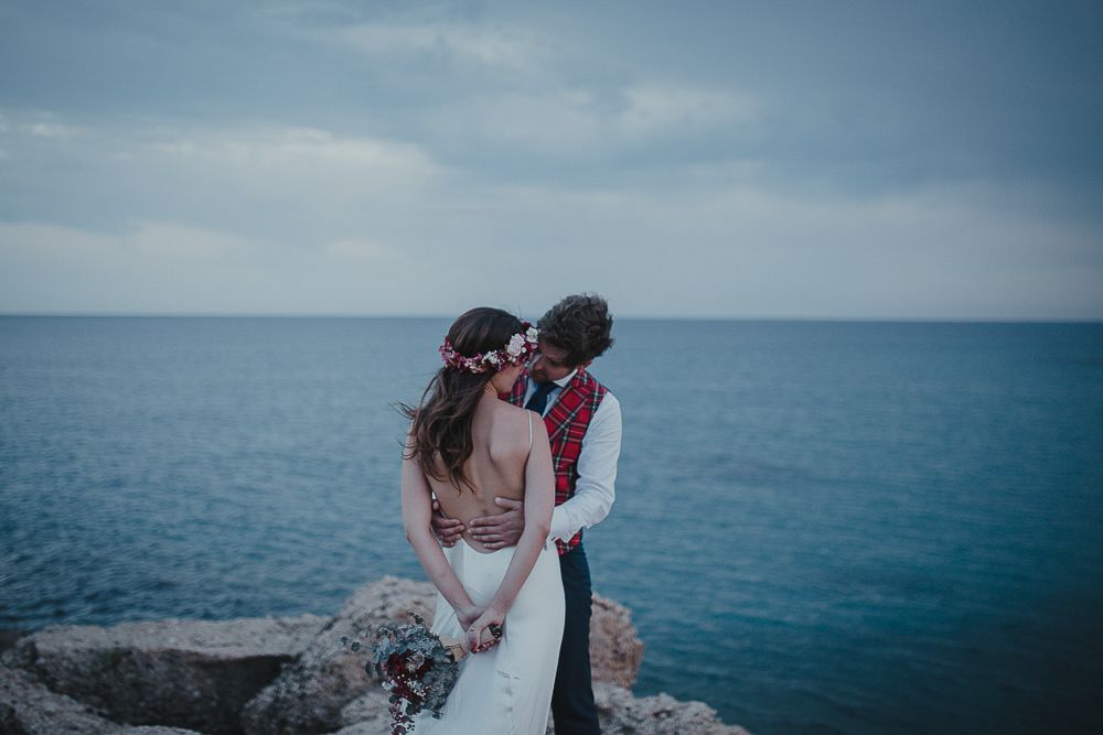 A couple in wedding attire embrace on coastal rocks overlooking bright blue waters.