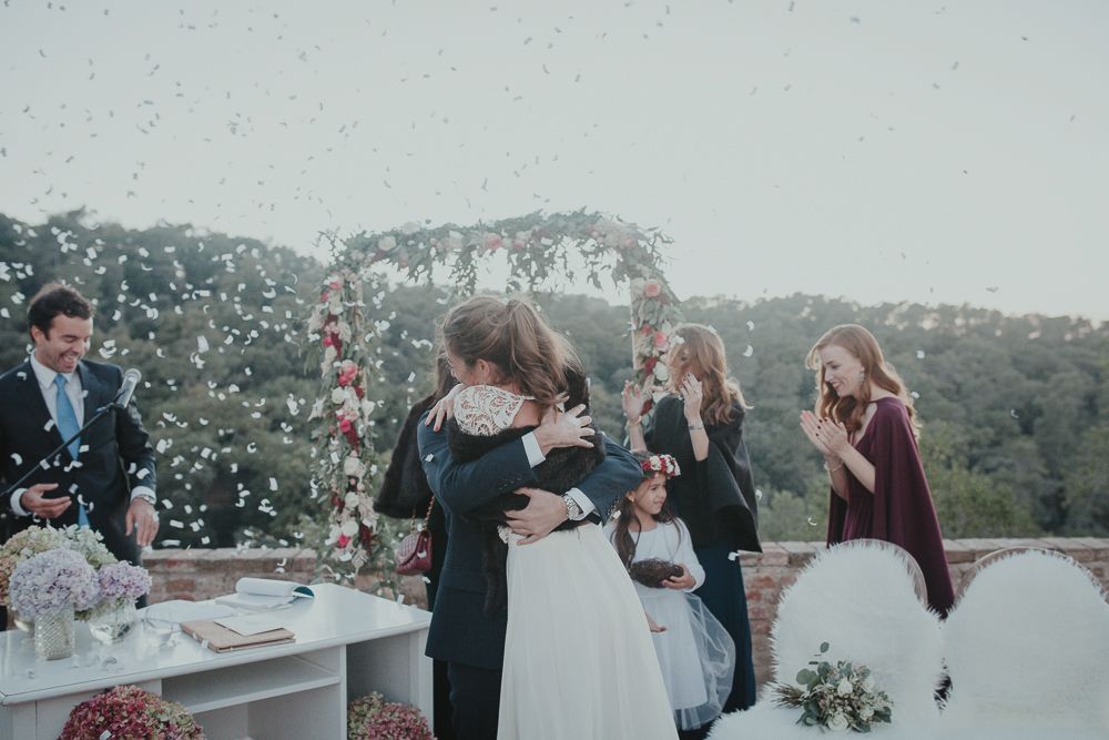 Wedding celebration outdoors with guests gathering around a white table amid mountainous scenery.