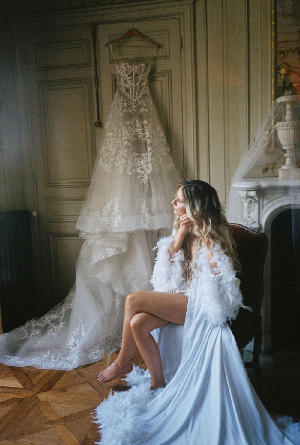 A romantic boudoir scene with a hanging wedding gown and feathered robe in an ornate room.