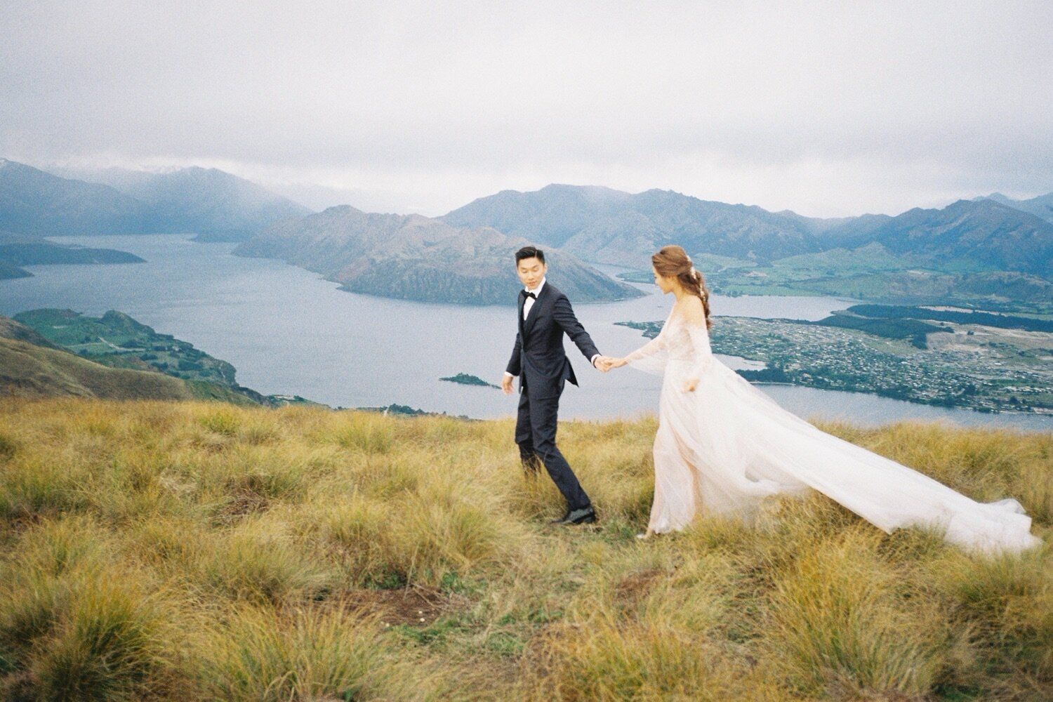 Wedding couple stands on mountain ridge overlooking scenic lake and hills in romantic outdoor shoot.