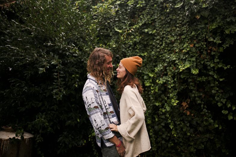 A couple in casual autumn wear poses intimately against green foliage.