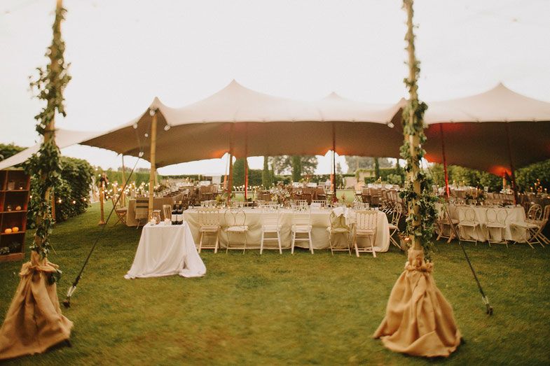 Elegant tent reception setup with draped fabric poles and white chairs on a lawn.