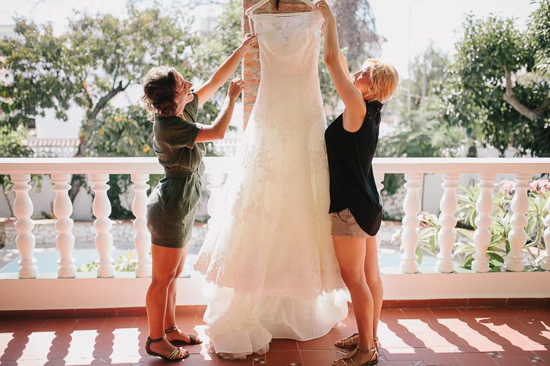 Wedding dress being held up by assistants on sunny balcony.