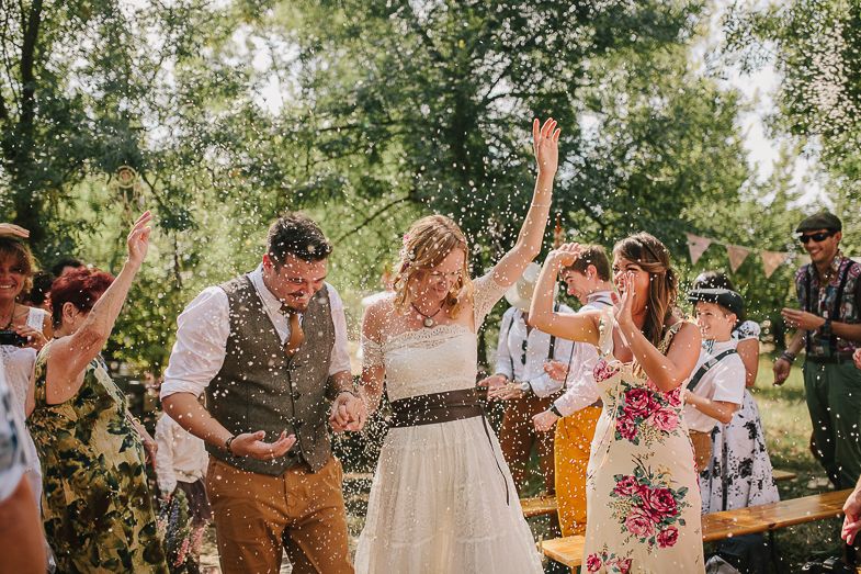 Wedding guests celebrating outdoors with bubbles and joyful dancing under green trees.