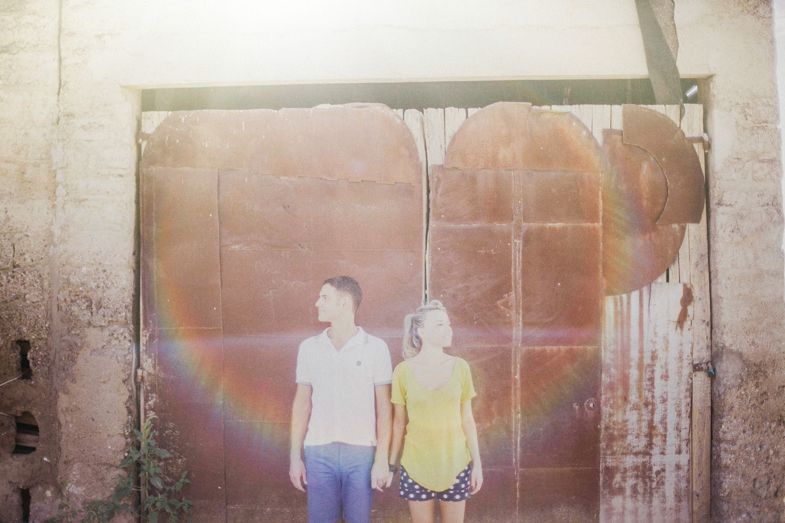 A couple stands against a weathered wall with a rainbow effect in the background.
