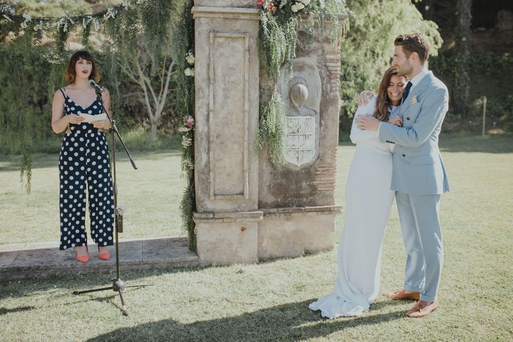 Couple in blue suit and white dress embrace during outdoor wedding ceremony while officiant in polka dot dress watches.