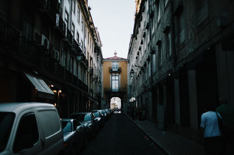 Narrow urban street with parked cars leading to an arched passageway.