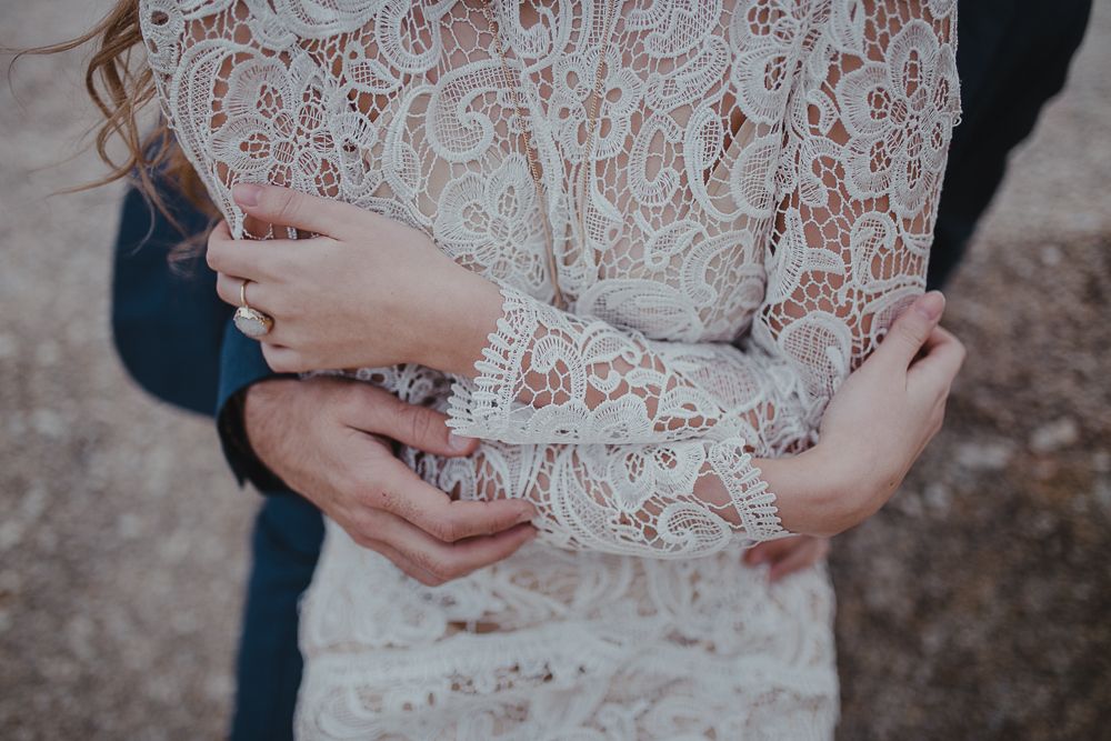 Close-up detail of delicate lace sleeves and embracing hands against a natural backdrop.