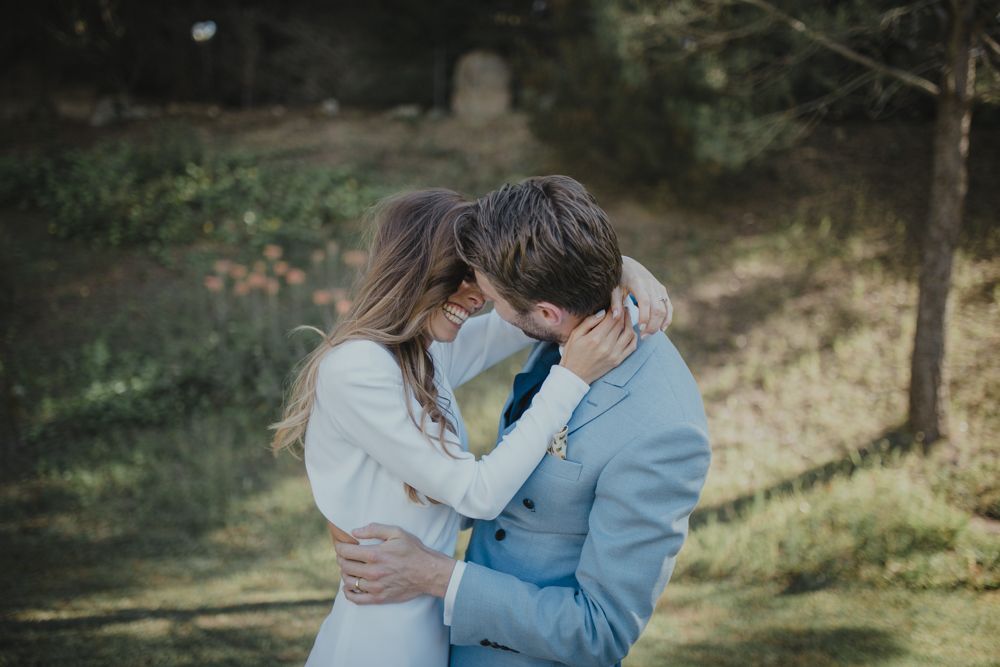 Romantic embrace between newlyweds in blue suit and white dress against natural backdrop.
