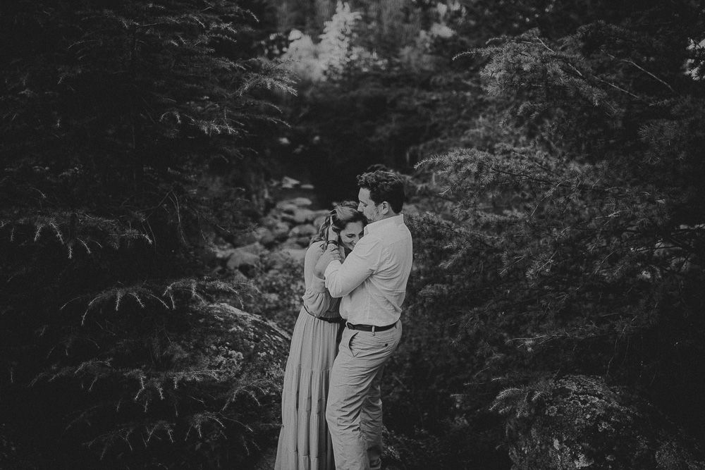 A romantic embrace among tall evergreen trees in dark moody lighting.