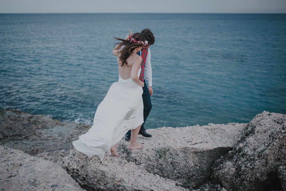 White dress flowing in wind as couple stands on rocky ocean overlook.