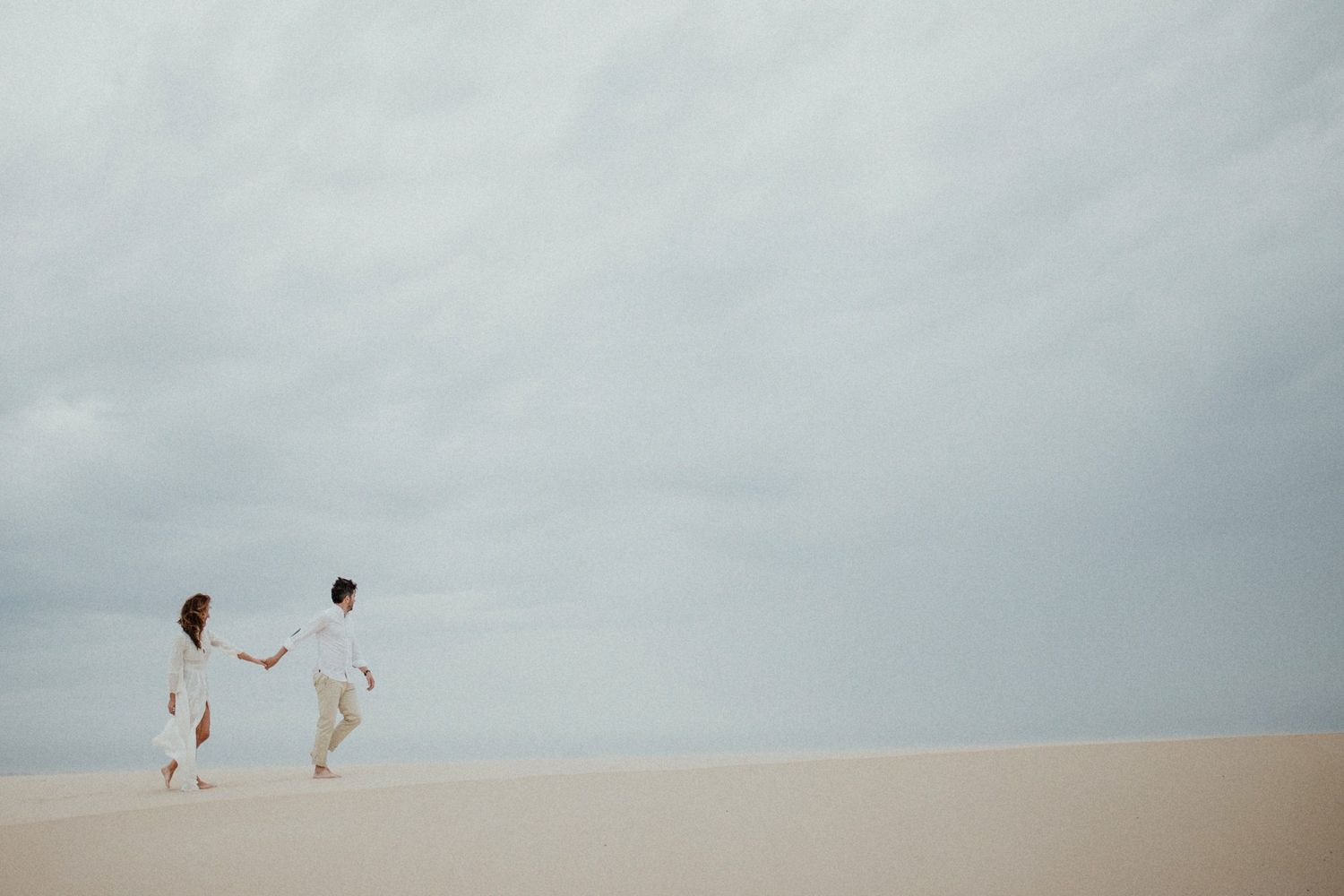 Distant view of two people walking along a minimalist beach landscape under cloudy skies.