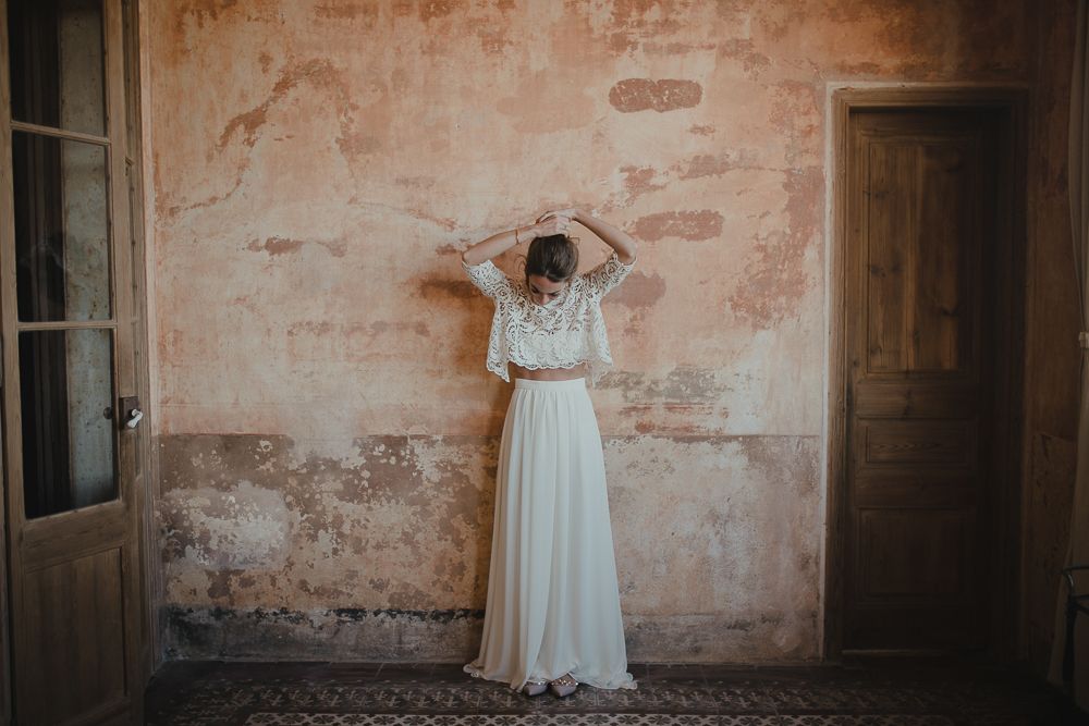 A two-piece white lace wedding dress shown against a weathered pink wall with vintage doors.