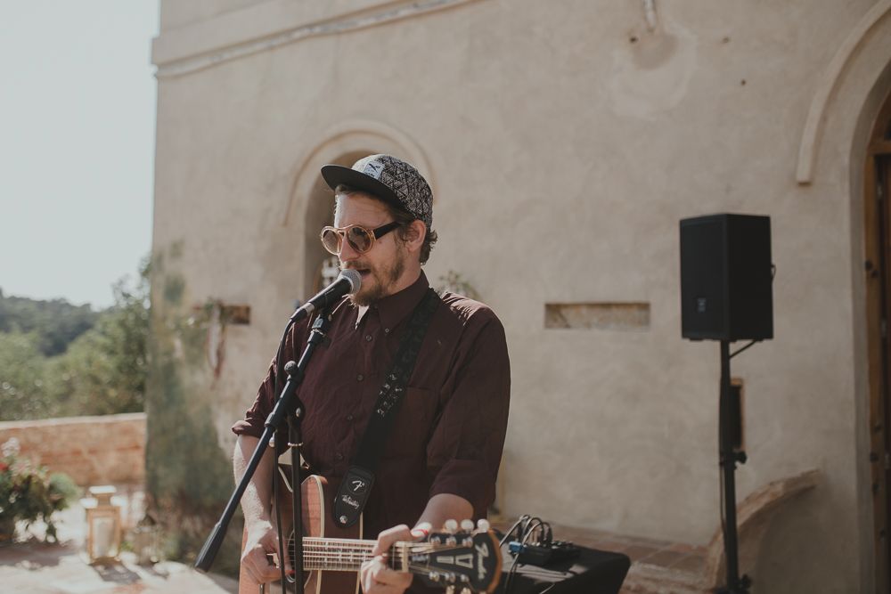 Musician performing with acoustic guitar in front of white stone building.