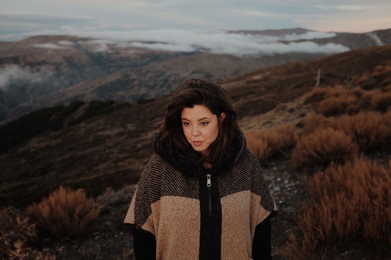 A person in a brown coat stands against a misty mountain landscape at sunset.