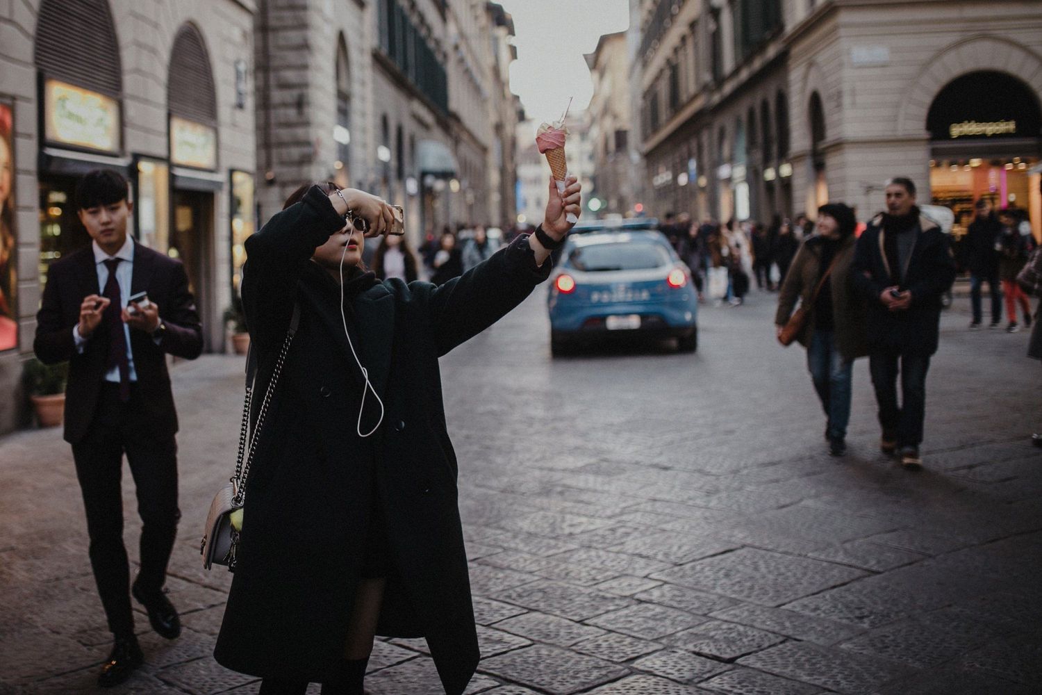 Person in black coat taking photo on busy European city street with historic architecture.