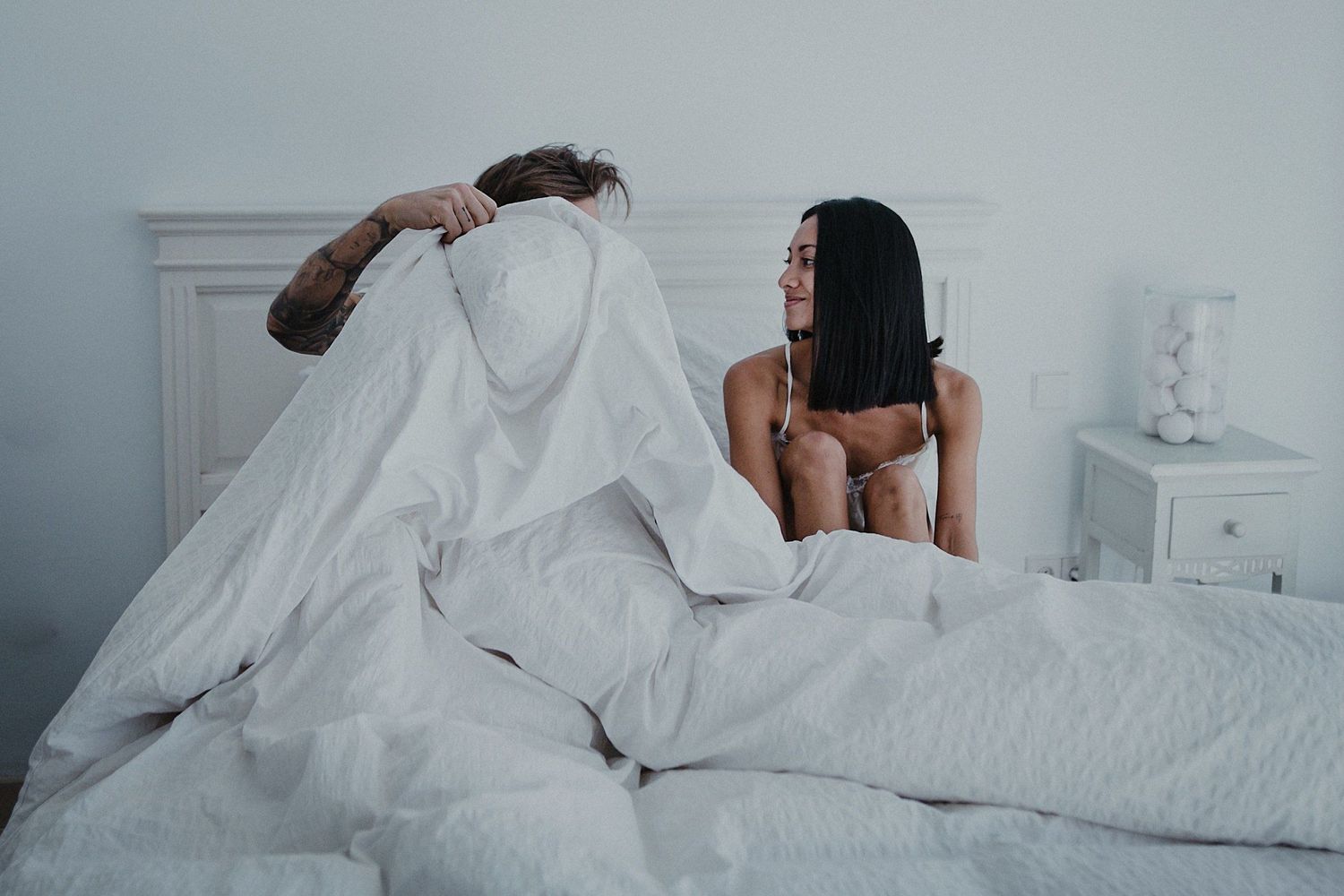 A couple playfully relaxing together on white bedding in a bright bedroom.