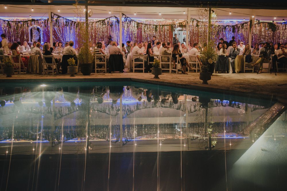 Poolside wedding reception with purple lighting and elegant white decor reflected in water.