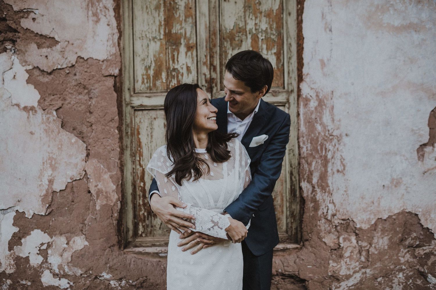 An intimate portrait of a couple in wedding attire against a weathered wall with peeling paint.