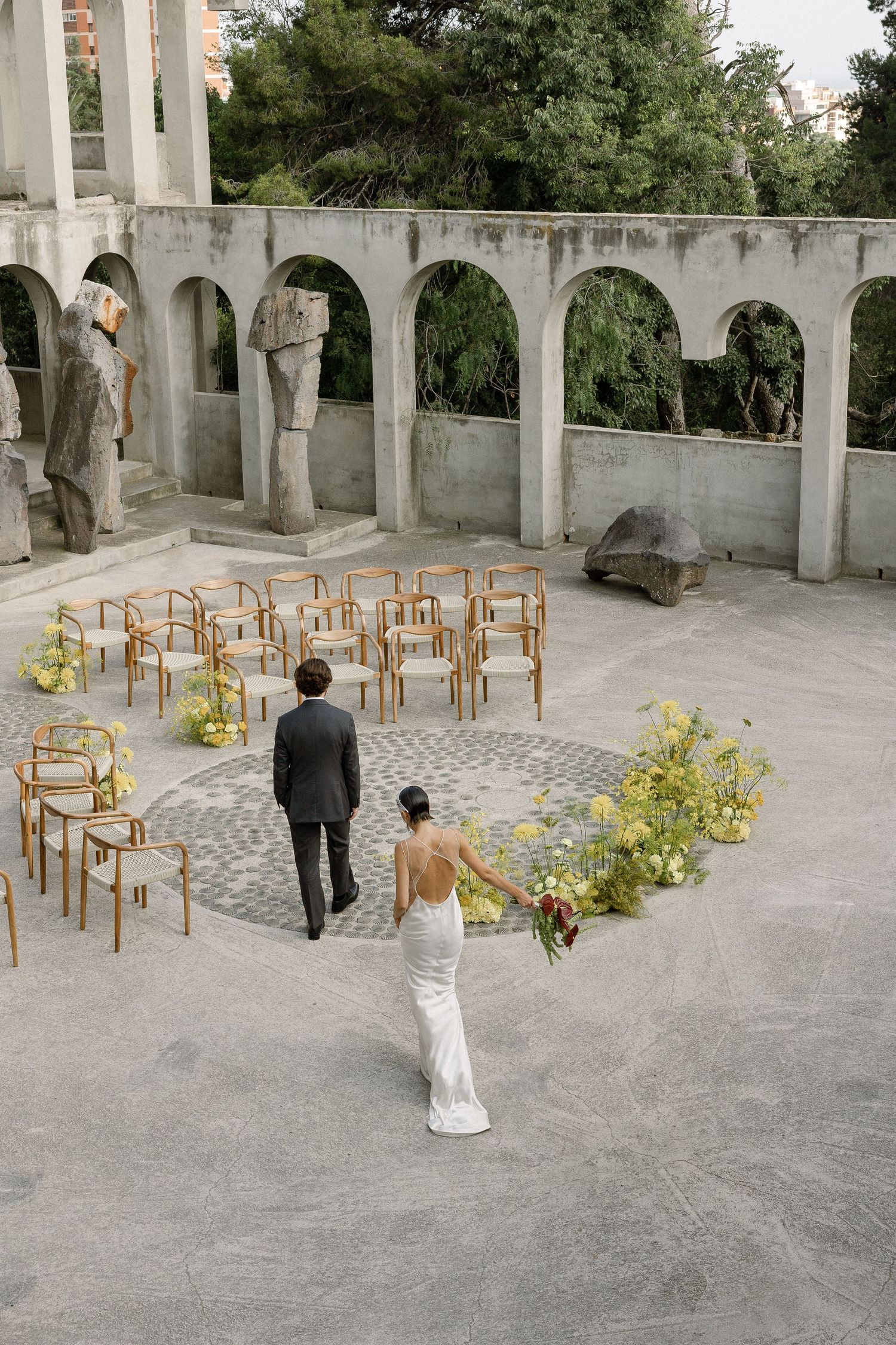 Couple walking through an outdoor stone colonnade with wooden chairs and yellow floral arrangements for a wedding ceremony.