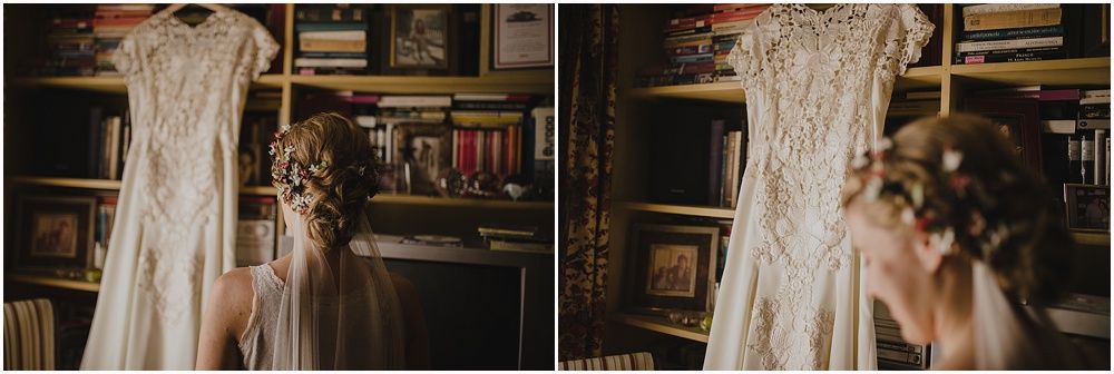 Elegant lace wedding dress hanging in front of vintage bookshelves with dried florals.