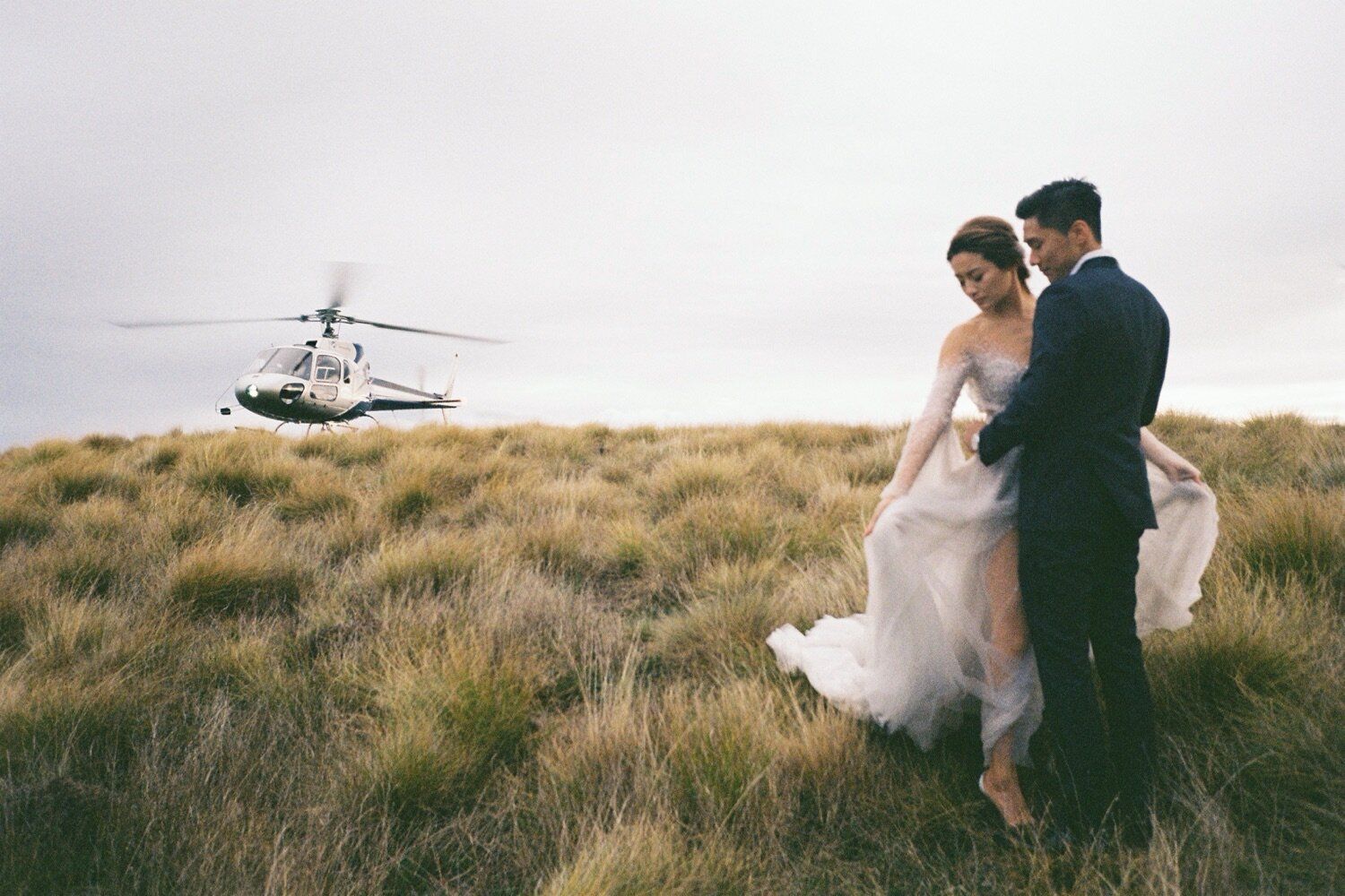 Wedding couple watches helicopter approach during mountaintop photo session.