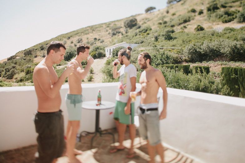 Group of shirtless people enjoying drinks on a sunny Mediterranean terrace with mountain views.