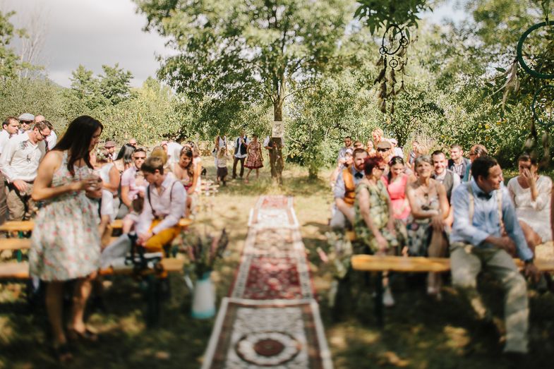 Outdoor wedding ceremony with guests seated on wooden benches under trees.