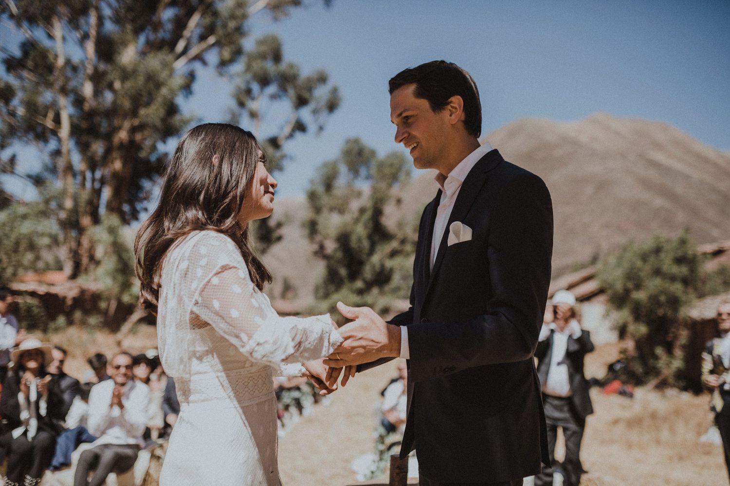 A couple holds hands during an outdoor wedding ceremony with mountain views and trees in the background.