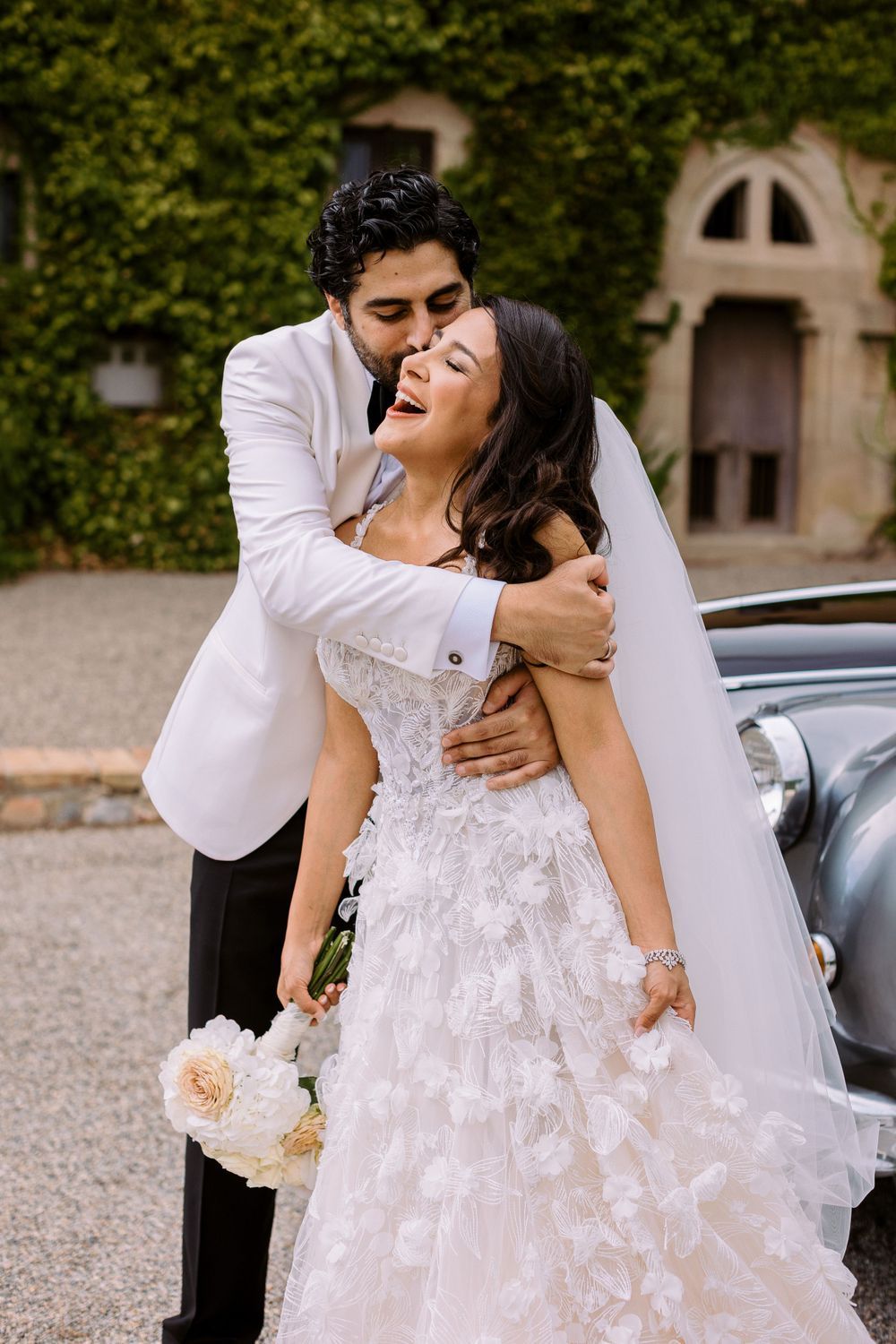 A couple shares romantic moments near a vintage car in formal wedding attire.