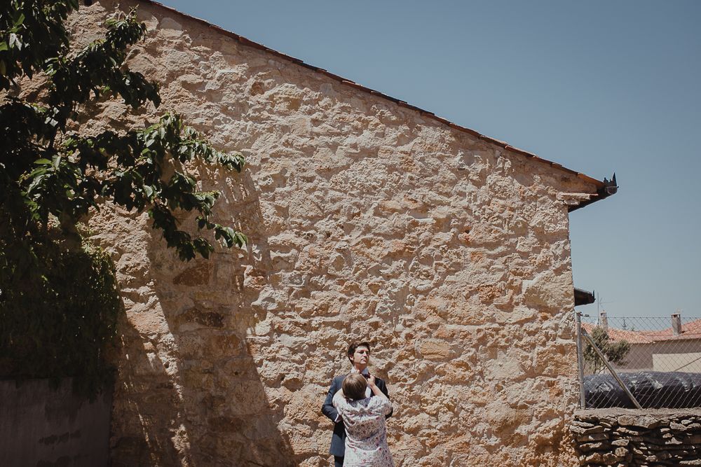 Figure in formal wear poses near corner of stone building under blue sky.