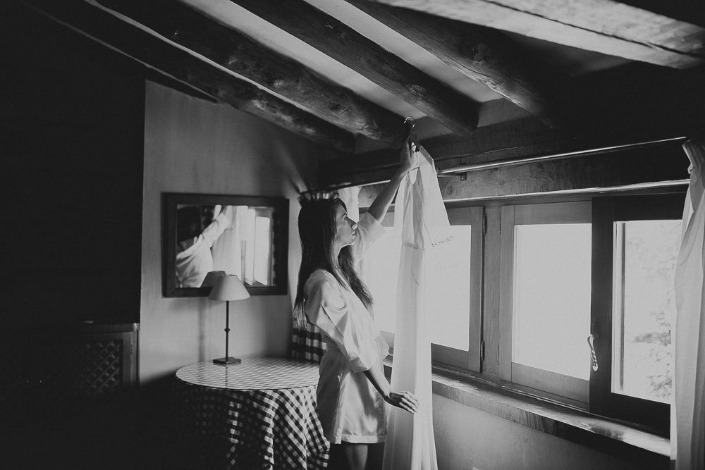 A white dress hangs from exposed wooden beams in a rustic room with windows.