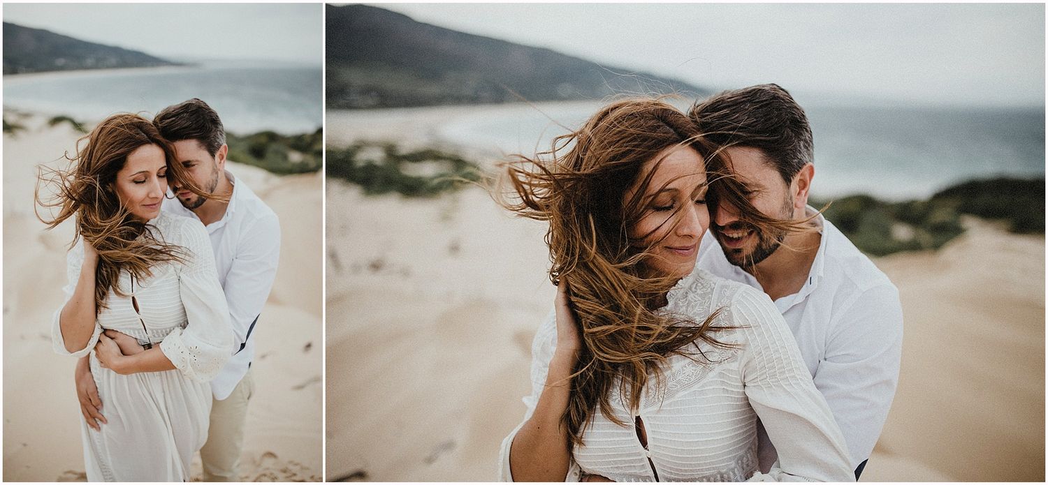 Wind-swept couple sharing laughter on sandy beach dunes by the ocean.