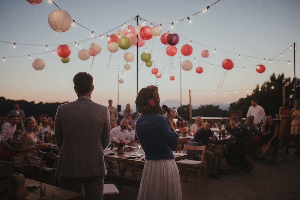 Outdoor wedding reception under string lights and pink paper lanterns at sunset.