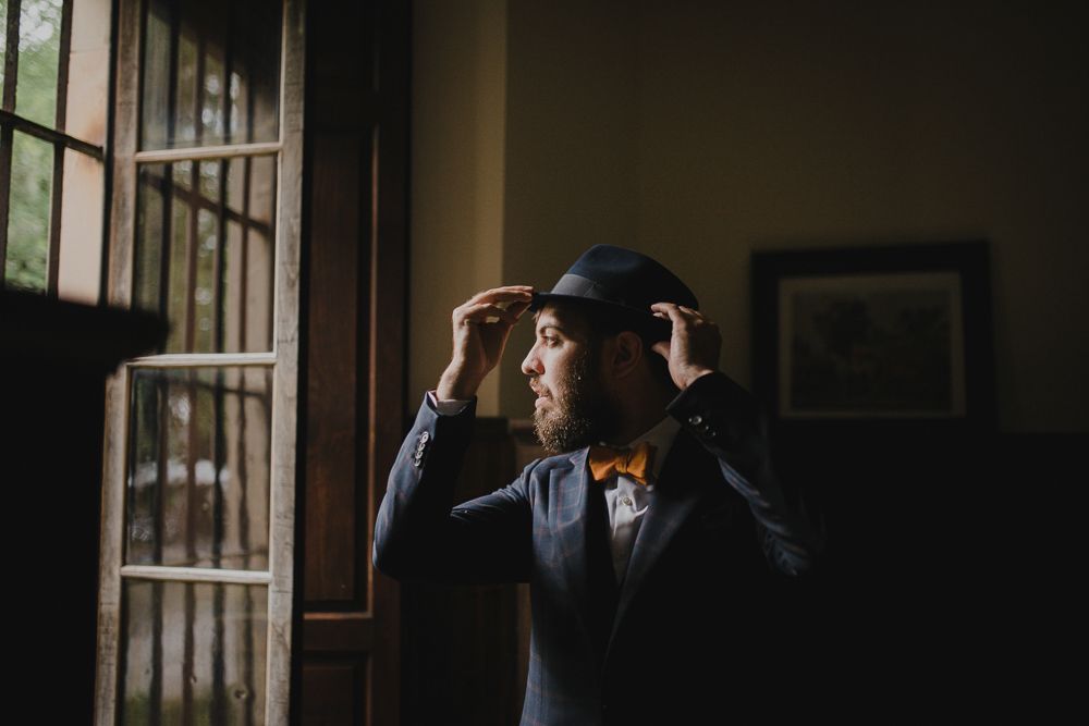 A groom in a dark suit and hat adjusts his attire while standing by a window in moody lighting.